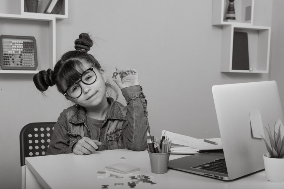 Young girl with glasses sat at a desk holding up the letter R. In front of her on the desk is paper, a laptop and a pot of colouring pencils. Image is for the head of the hiring a product manager article.