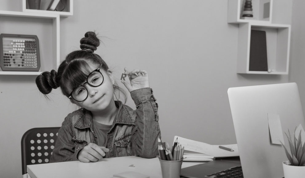Young girl with glasses sat at a desk holding up the letter R. In front of her on the desk is paper, a laptop and a pot of colouring pencils. Image is for the head of the hiring a product manager article.