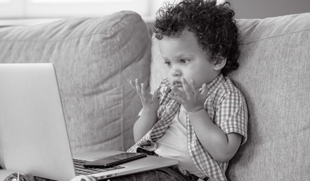 black and white image of a child sat on the sofa with a laptop on his lap. The child is looking confused (probably because he's unsure on whether to switch to a career in tech)