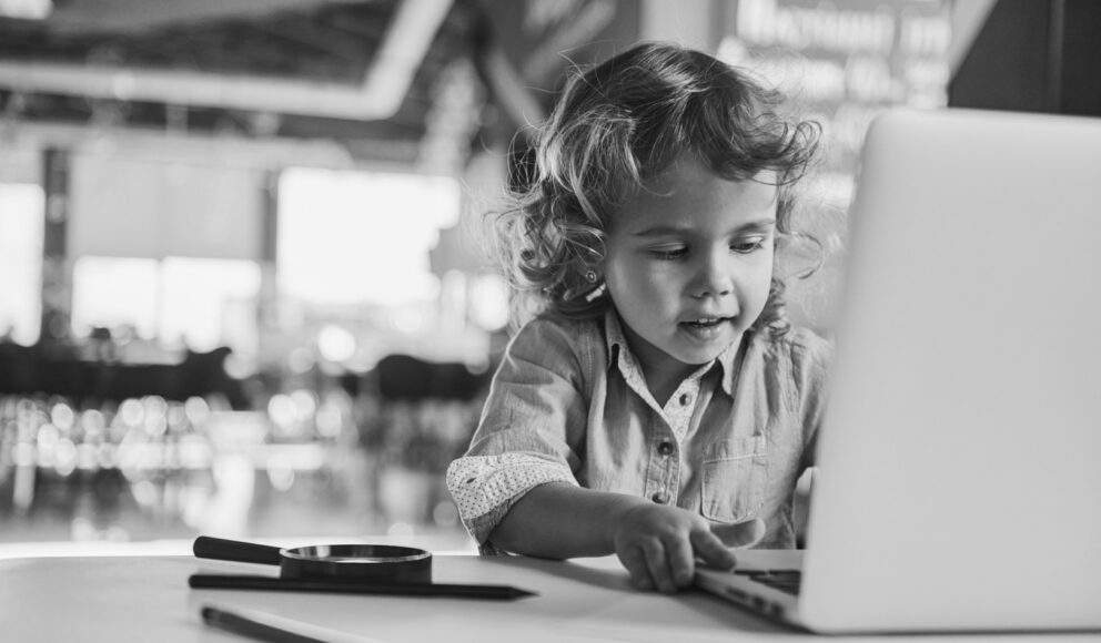 Small child sat at a desk on a silver computer. Image is in black and white. Image for the 'top 5 reasons to learn java' blog.
