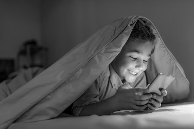 Black and white photo of Kid boy playing smartphone lying on a bed at night. 7 years old child boy using smartphone at night in bed. Boy in bed texting on smartphone. Child with smart phone. Cover image for the tech candidates blog.