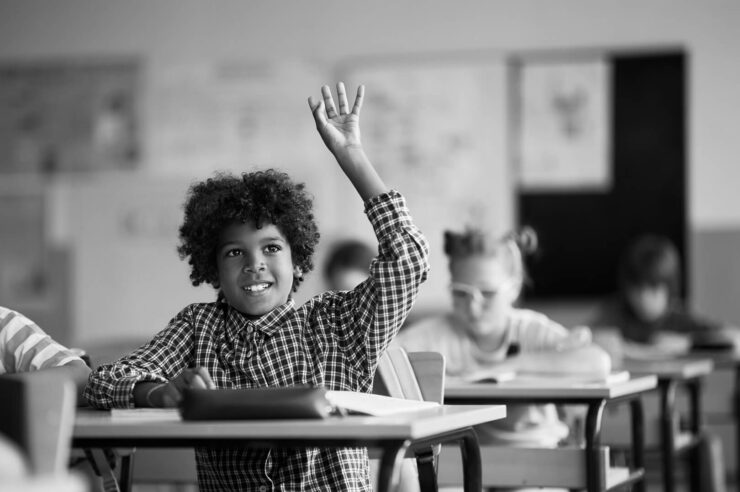 Black and white image of a child sitting at his desk in a classroom. He has his hand up to ask a question. Image is a cover image for the 5 questions to ask at the end of an interview to wow tech employers blog.