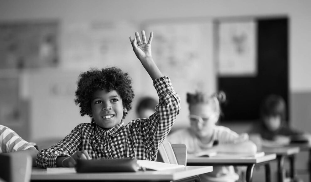 Black and white image of a child sitting at his desk in a classroom. He has his hand up to ask a question. Image is a cover image for the 5 questions to ask at the end of an interview to wow tech employers blog.
