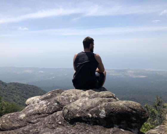 Man sitting on top of a mountain, facing away into the horizon.