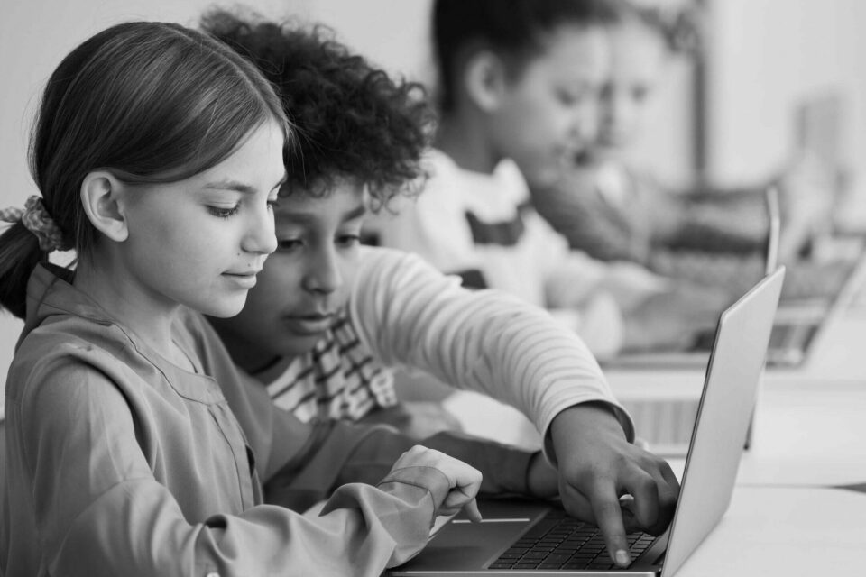 Black and white image of two kids sitting at a desk pointing at a laptop. Image is for the how to retain tech talent blog image.
