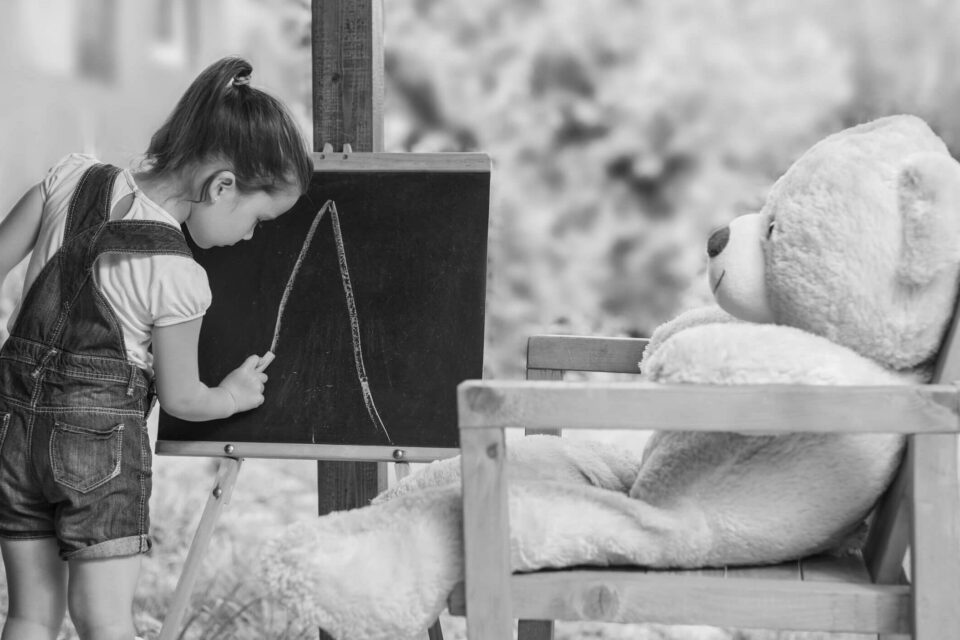 Black and white image of a girl drawing on a chalk board, with her large teddy sat next to her. Cover image for the Software Engineer interview process blog.
